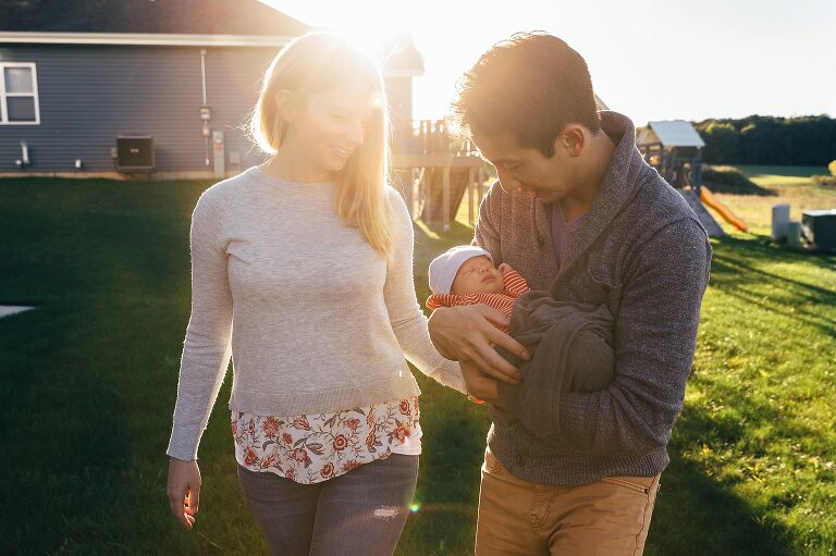Mom and Dad walk through sunshine backyard holding newborn