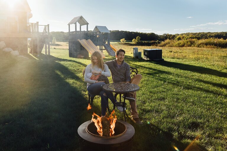 Mother and father sit in the backyard on a sunny day holding newborn in front of a firepit. 