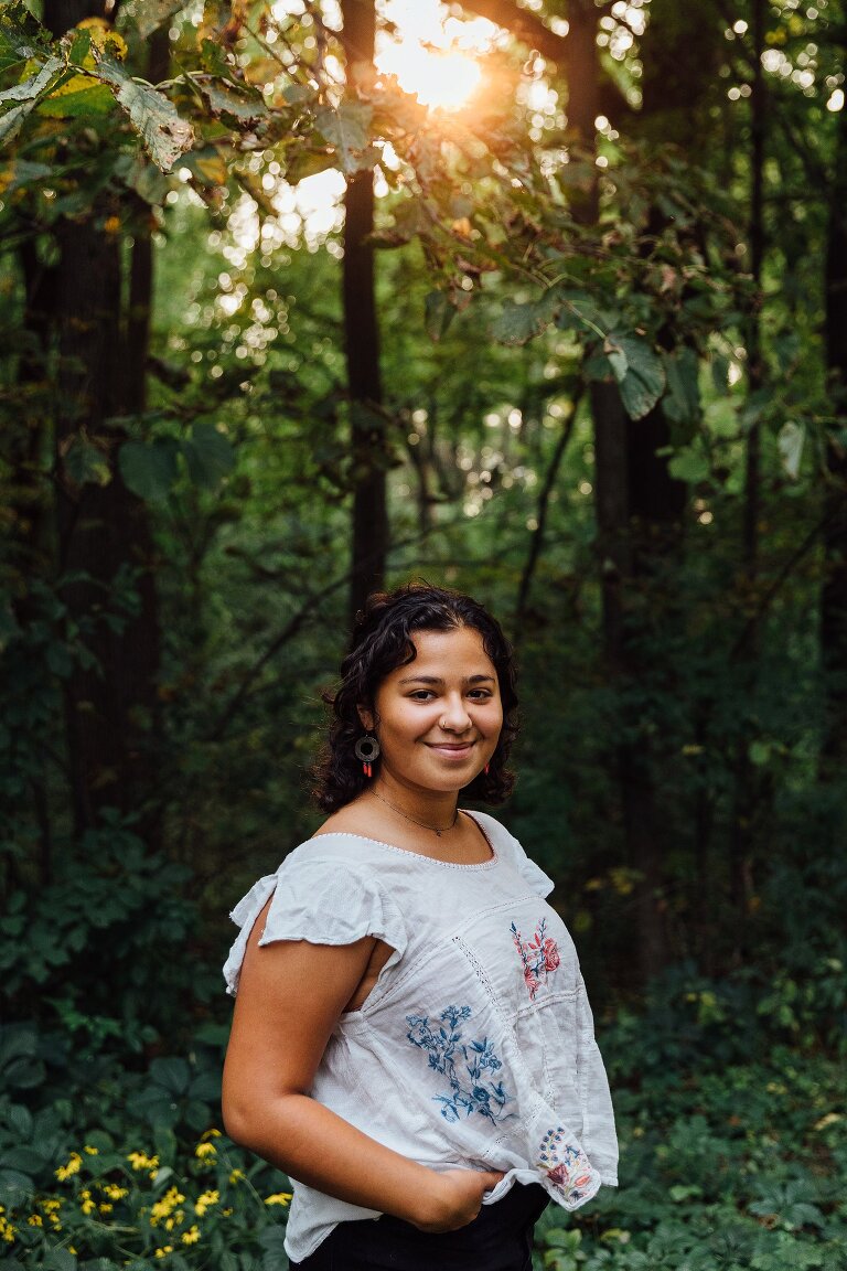 High school senior portrait, smiling and standing in a forest with dappled light 