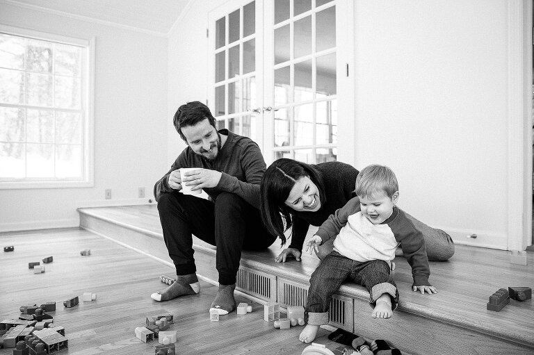 Black and white. Dad drinks coffee and smiles as mom and young son smile in a playroom. 