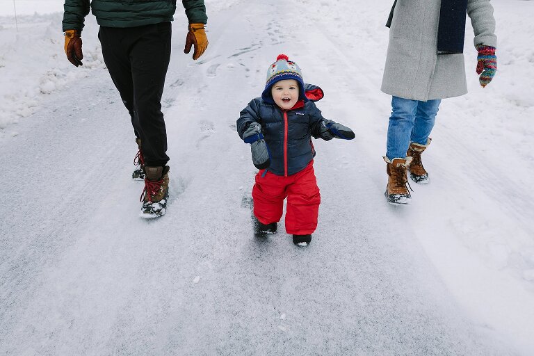 Toddler boy walks down street smiling making snowy footprints. Two parents walk beside him. 