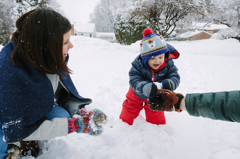 Young toddler smiles as he grabs a snowball . Mom looks on happy. 