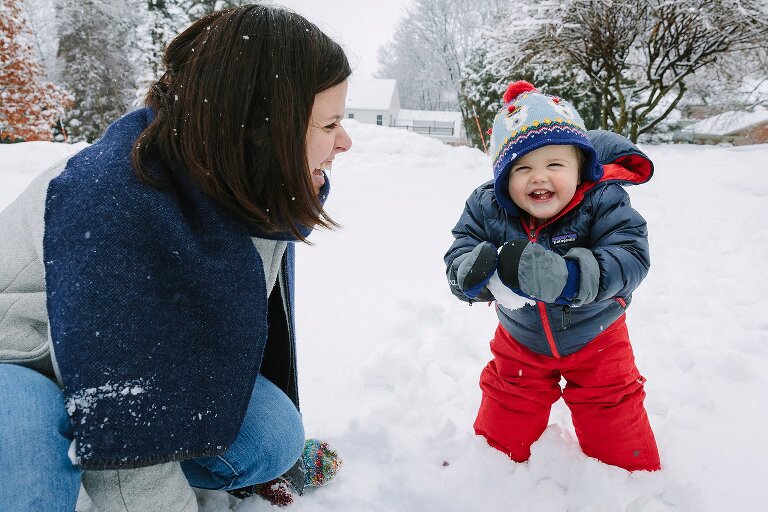 Young toddler smiles as he holds a snowball. Mom looks on happy. 