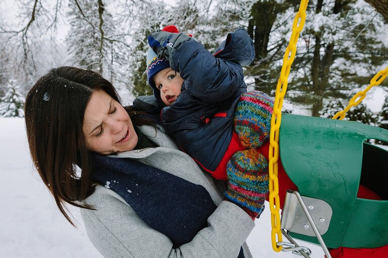 Mom helps toddler out of a swing, which is cumbersome with all the winter gear on.