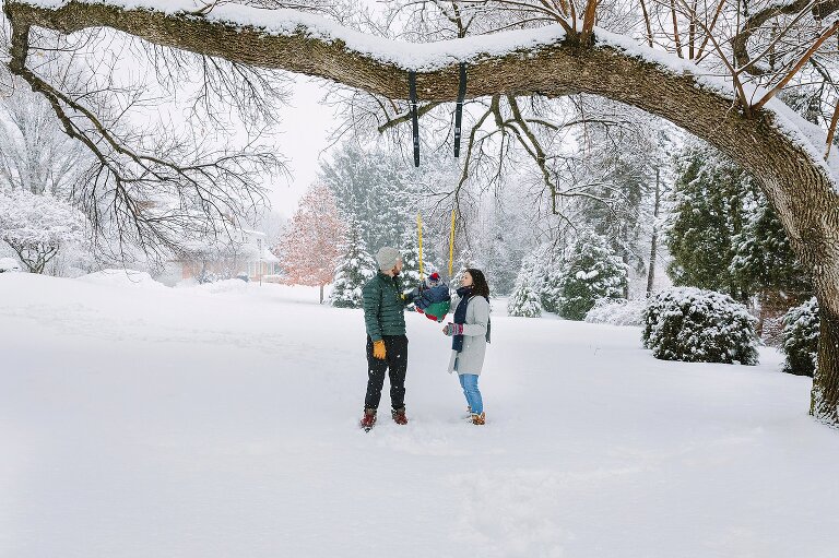 Mom, Dad, and toddler swing from a tree in the backyard on a snowy day