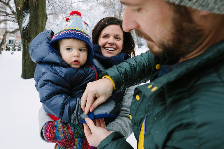 Mom holds toddler while Dad puts mittens on toddler in the snowy day