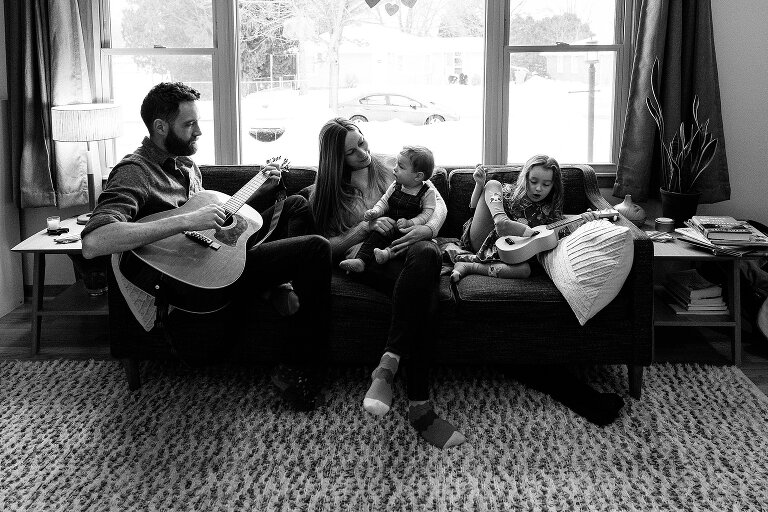 Black and white. Family sits on sofa and listens to father play guitar. Girl sturrums alng on ukulele. 