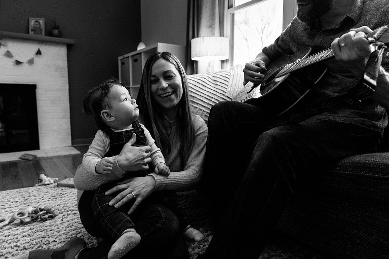 Black and white. Infant held by mother watches as father plays guitar. 