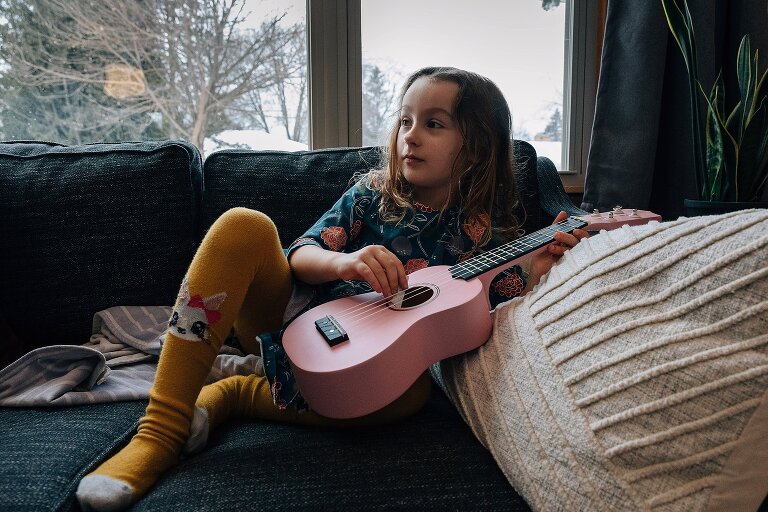 Girl sits on sofa holding pale pink ukulele. 