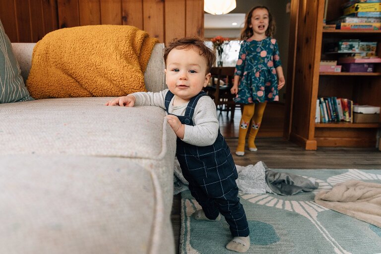 Infant stands supported by sofa while girl laughs in the background.