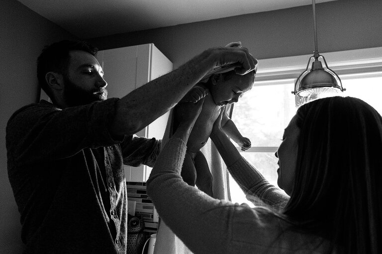 Black and white. Mother holds infant still while father puts a hat on the infant's head
