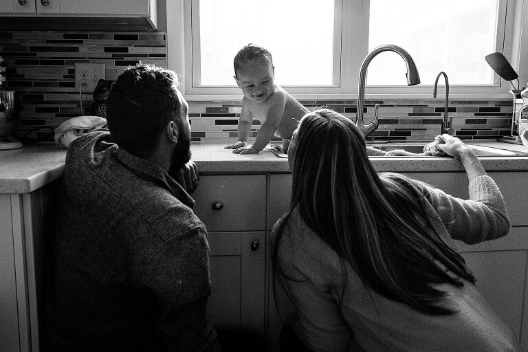 Black and white. Mother and father give smiling infant a sink bath. 