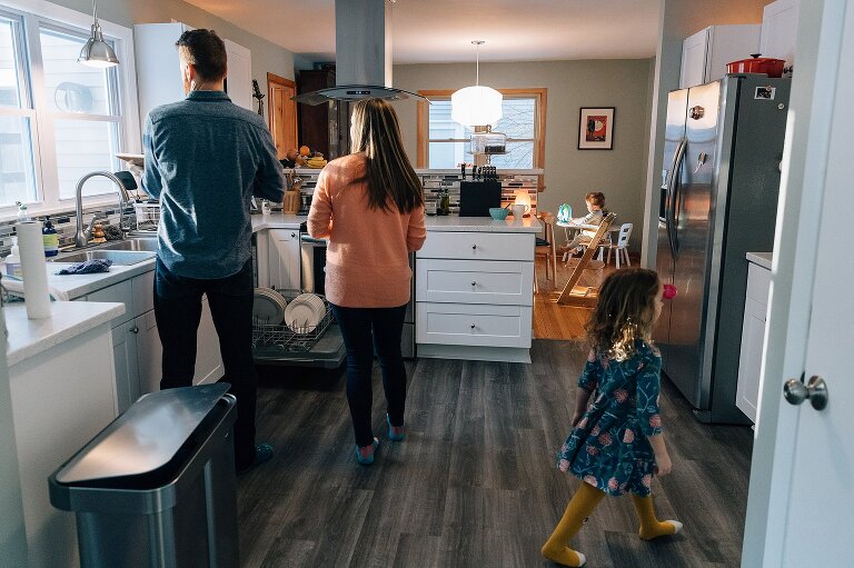 Parents clean dishes while girl walks across kitchen, infant sits in highchair in the dining room.