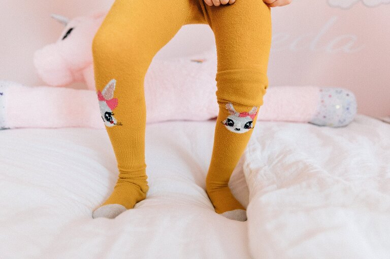 Girl pulls up mustard yellow tights with a bunny on the knees while standing on pale pink bed with pale pink walls 