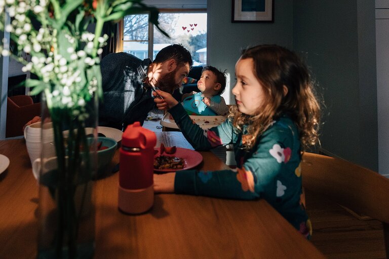 Sunlight on girl in front at the dining room table, but focus on dad and infant son behind her. 