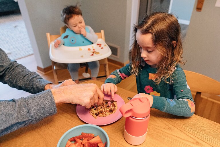 Dad helps daughter cut winter sunday pancakes while infant eats at high chair behind them.
