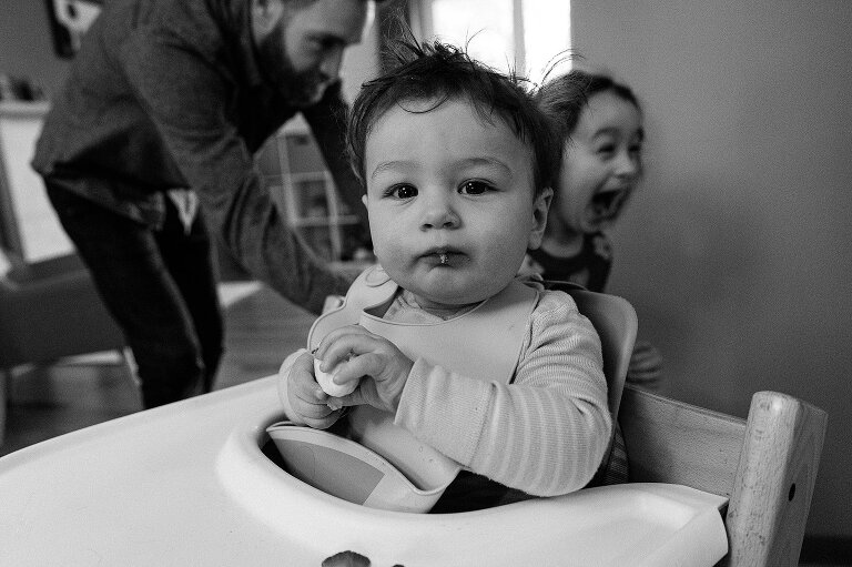 Black and white. Environmental portrait of infant boy with dad and joyful sister run through the background