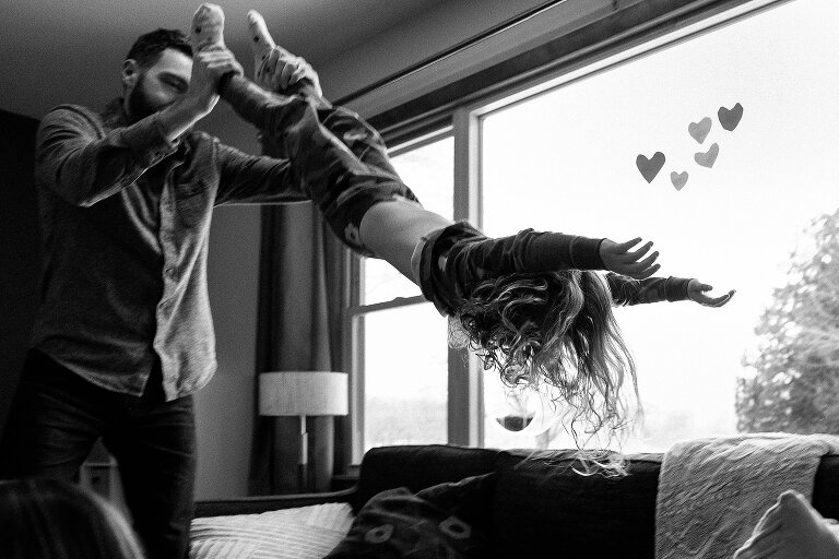 Black and white. Dad holds daughter by ankles and playfully swings her. Construction paper hearts on the window.