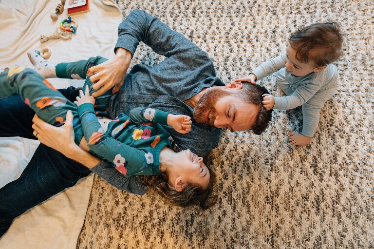 Dad lays on floor holding daughter while infant son pulls dad's hair
