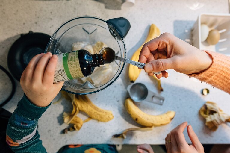 View over blender of young girl pouring vanilla into teaspoon held by adult 