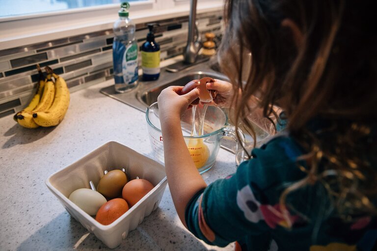 Girl cracks egg into a measuring cup. Bananas sit on kitchen counter. 