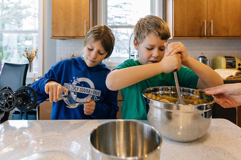 Hand on far right holds bowl while boy stirs the bowl and girl carefully pours a tablespoon of lemon juice