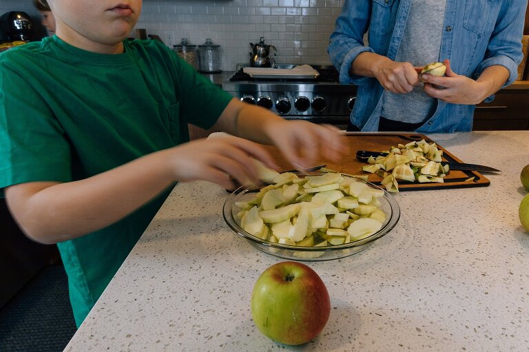 Motion blur shows boy tossing apples into pie plate while mom continues to cut apples in background making Favorite Apple Pie