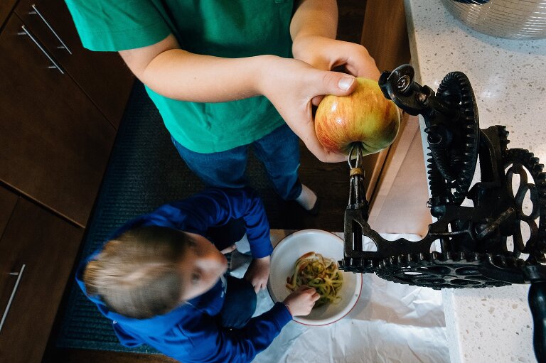 boy holds onto apple on old-fashioned clamp peeling machine while girl waits for peels below 