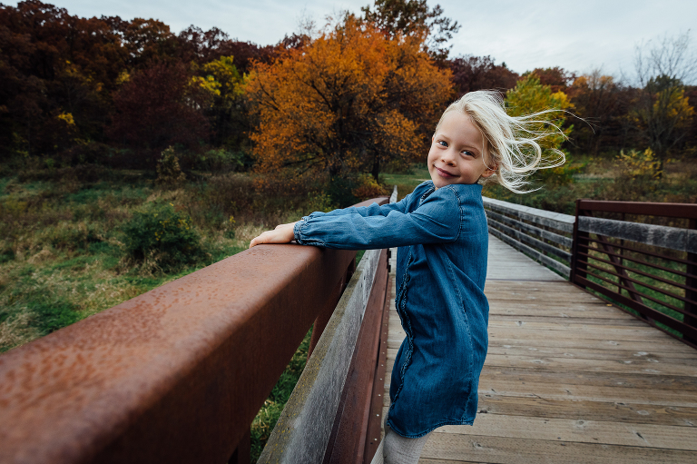 camera aware girl pulled up on bridge railing, wind blowing hair back, autumn forest in background 