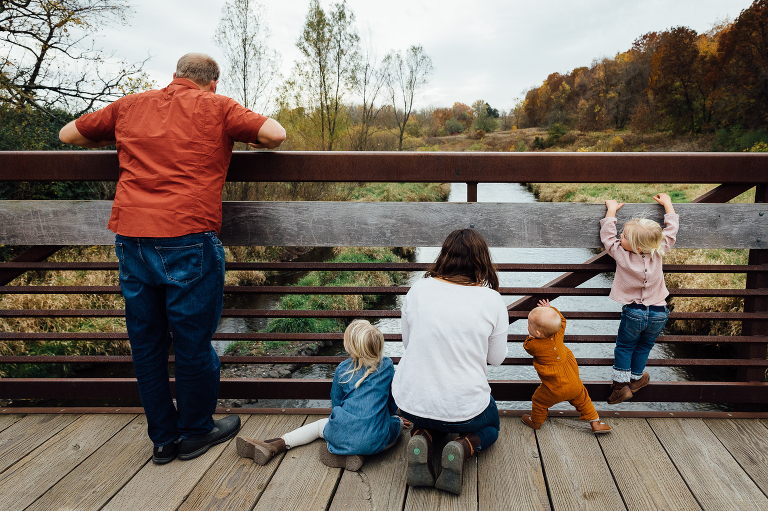 family of five stand or sit on an iron and wooden bridge overlooking a small river with autumn forest in the background
