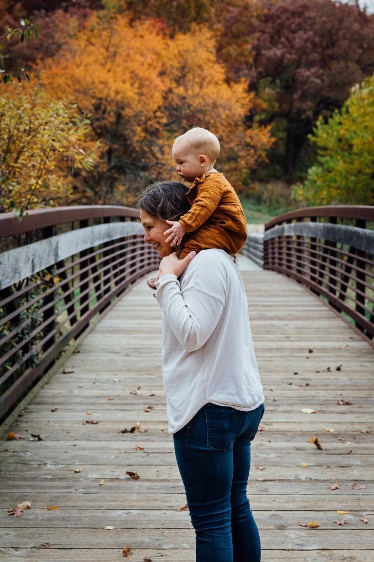 mom with baby on shoulders stands on a bridge in an autumn forest 