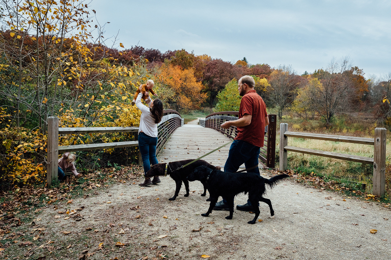 mom holds baby to the sky while dad approach with two dogs on a leash in an autumn forest