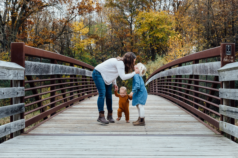 mom holds baby's hand with one hand and touches older daughter on the face with other for a kiss in an autumn forest 
