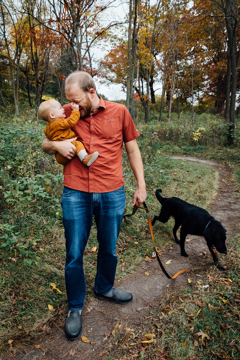 baby playfully squeezes dad's nose; dad holds baby in one hand and dog leash in another 