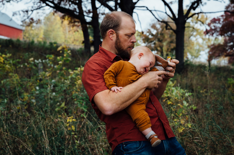 dad hilariously holds baby in awkward way as he keeps her safe and tries to open a snack at the same time. Baby smiles, autumn forest and red barn in background