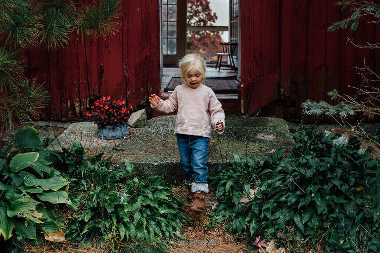 girl walks down step away from a dark red barn with windows to trees on the other side