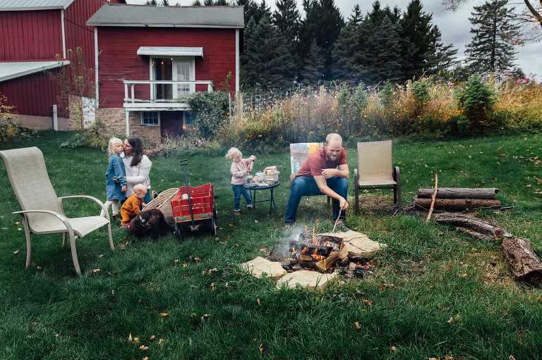 family of five prepare a fire in the background, garden and red barn visible