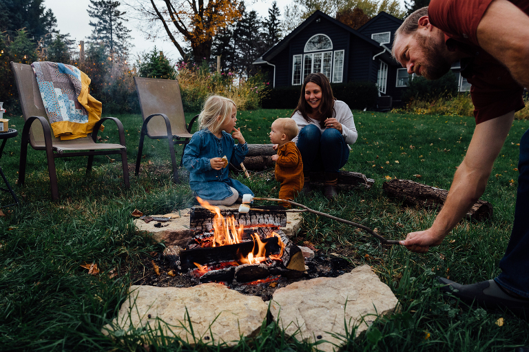 older girl points to her mouth with s'mores in it to the baby; dad in foreground roasting marshmallows