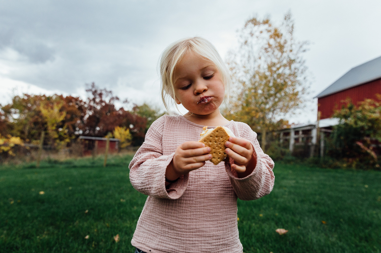 girl contemplatively examines s'mores in her hand with marshmallow on her face and autumn forest in the background 