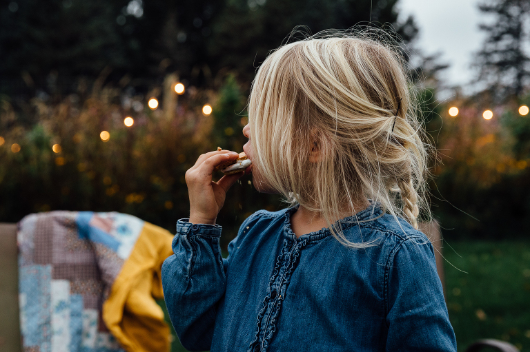 side view of girl eating s'mores at dusk with fairy garden lights in the background 