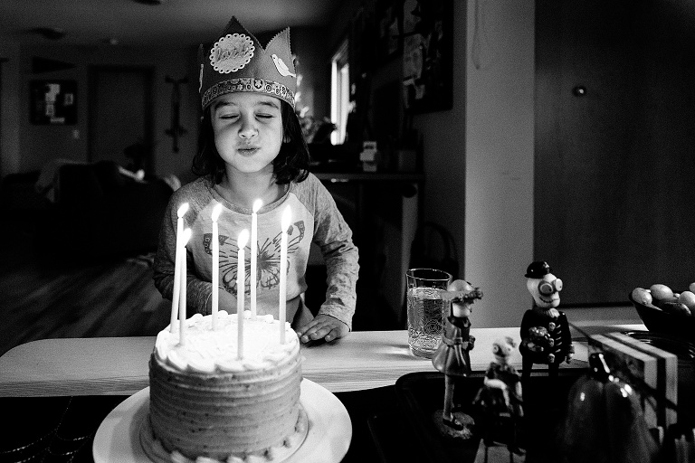 girl closes eyes, puffs cheeks, holds mouth closed as she hovers over a lit birthday cake