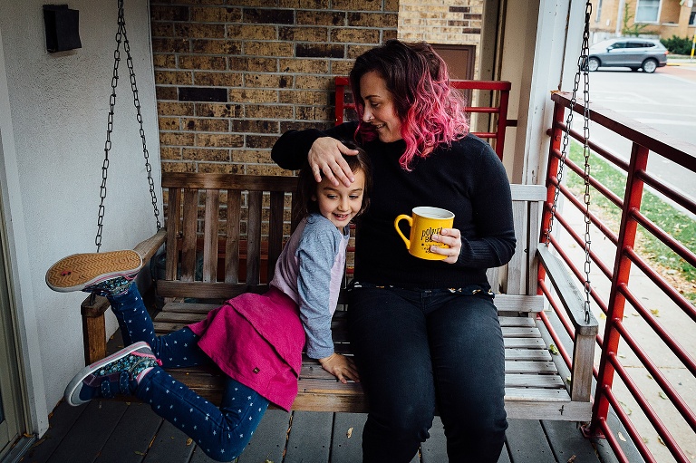 mom touches daughter's hair while holding a yellow cup of coffee and sitting on a porch swing
