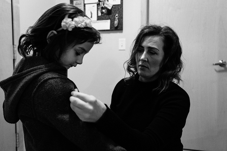 black and white. Mom helps daughter zip her coat in hallway light. 