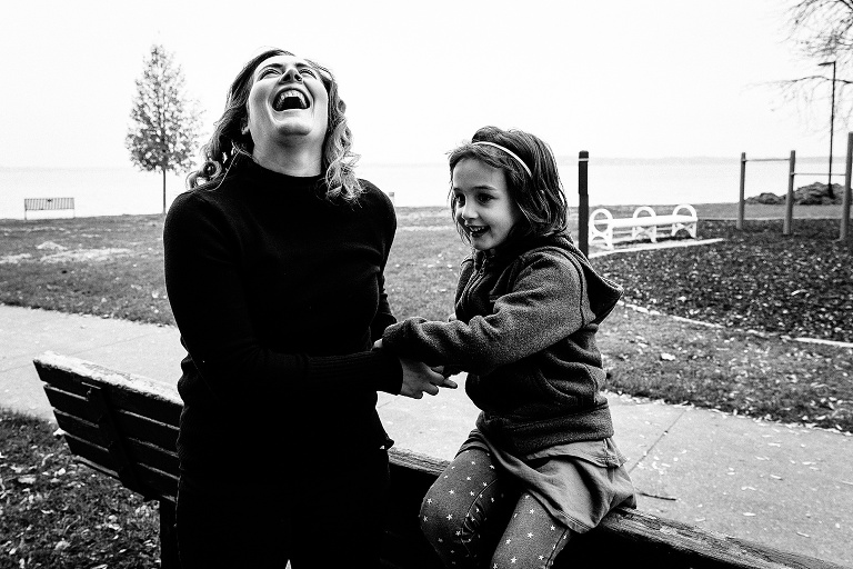 black and white. mom and daughter laugh at the park with lake behind them