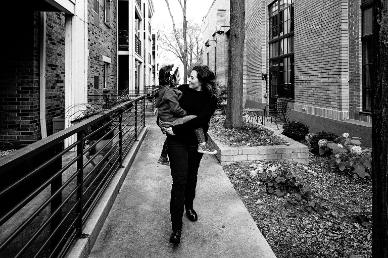 black and white. Mom carries girl through an outside building corridor. They look at each other while mom takes long strides. 