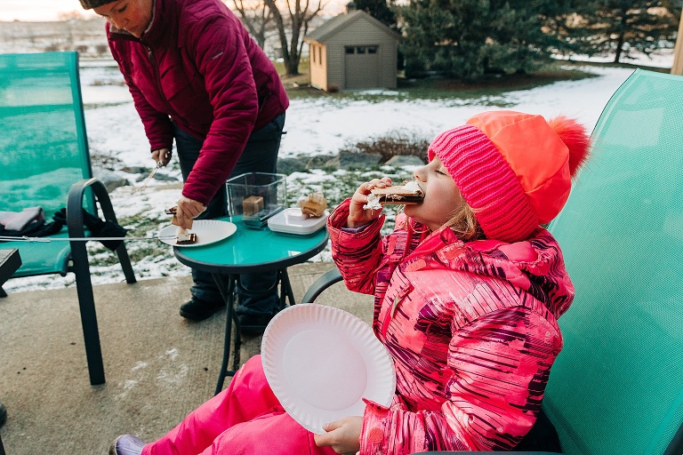 girl wears bright pink snow pants and jacket while eating a s'more at sunset in winter