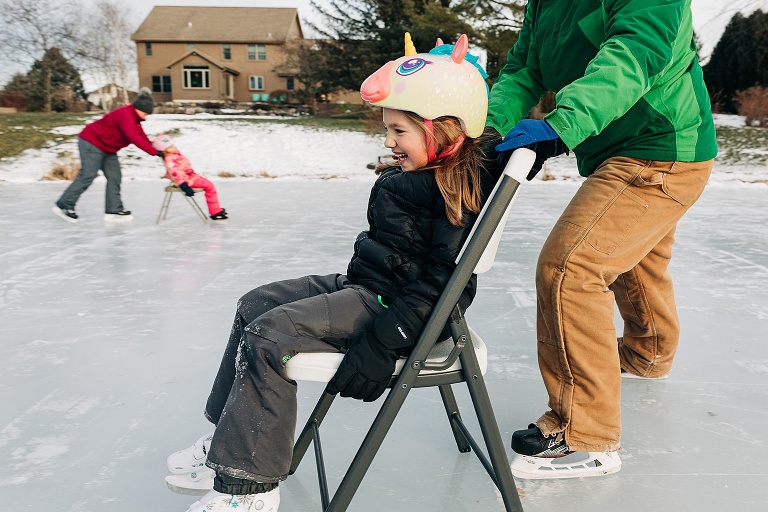 young girl sits in a chair on the pond ice while her father pushes her while ice skating