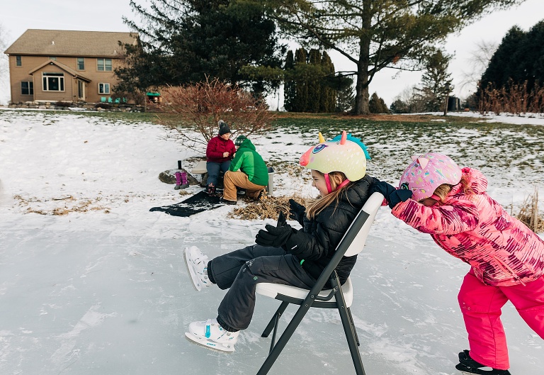 young girl pushes older sister on a chair on the ice with parents getting on their ice skates in the background 