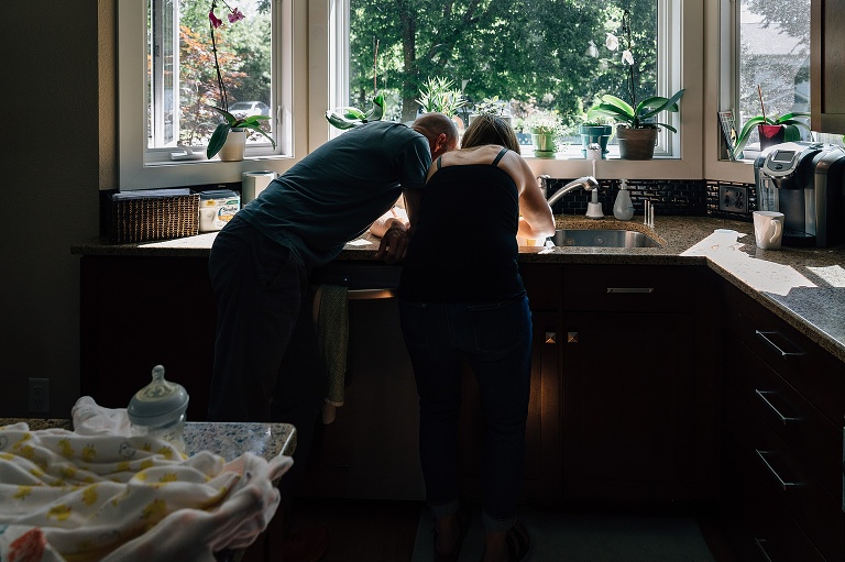 two parents lean over kitchen sink in sunlit room 