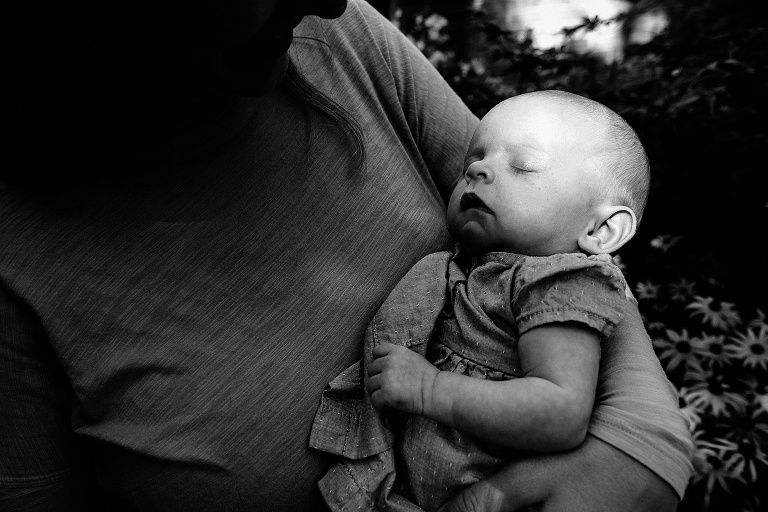baby sleeps in mother's arm outside, flowers and trees in background; black and white